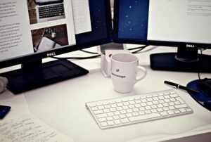 white ceramic mug between apple magic keyboard and two flat screen computer monitors